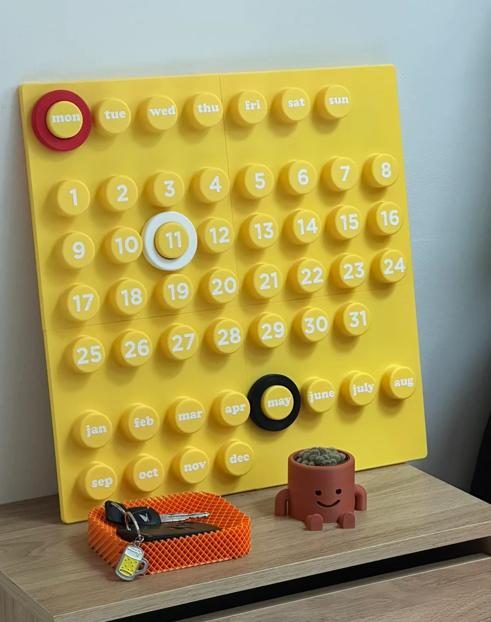 Calendrier perpétuel jaune à pastilles (jours, dates, mois), un vide-poche orange et un petit pot souriant avec un cactus