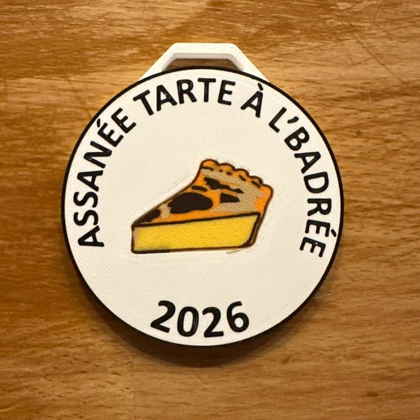 Médaille ronde imprimée, texte en relief « Assanée tarte à l'badrée 2026 » et une part de tarte en couleur